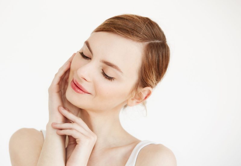 Portrait of young beautiful girl smiling with closed eyes touching face over white background. Facial treatment. Beauty cosmetology and skincare. Copy space.